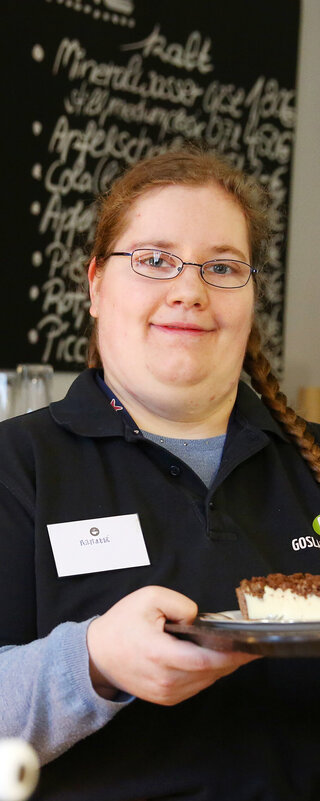 Eine Mitarbeiterin der Goslarschen Höfe steht im Café. Sie hält ein Tablett mit einem Kuchen und einem Kaffee in der Hand und schaut freundlich in die Kamera.
