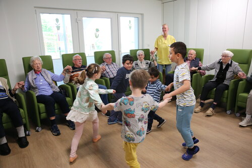 Kleine Kinder tanzen in einem Kreis, um sie rum sitzen ältere Menschen und schauen ihnen zu