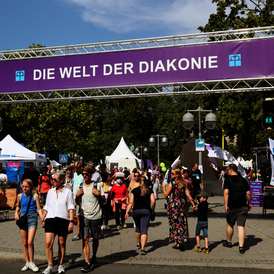 Menschen beim Entdeckertag. Sie laufen unter dem Banner "Die Welt der Diakonie" hindurch