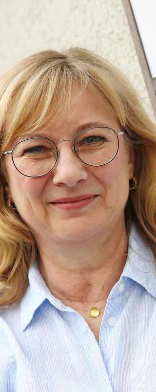Bettina Schmidtmann steht neben einem Schild der Einrichtung SINA. Sie schaut freundlich in die Kamera