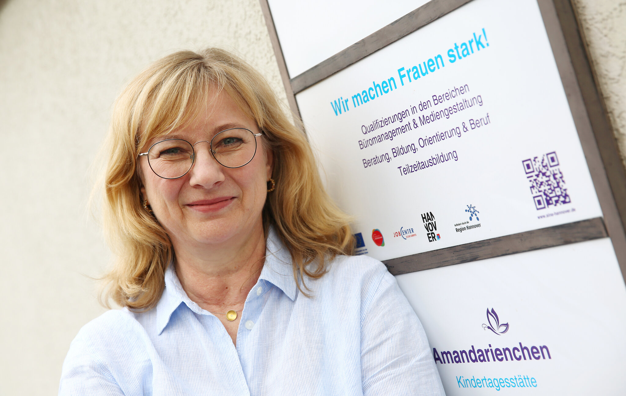 Bettina Schmidtmann steht neben einem Schild der Einrichtung SINA. Sie schaut freundlich in die Kamera
