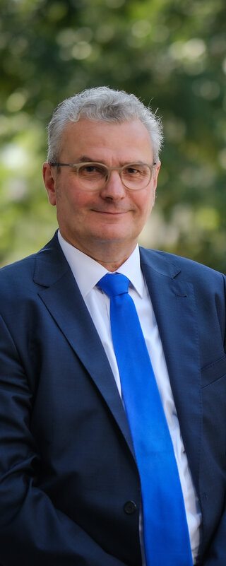 Herr Lenke und Frau Timm stehen nebeneinander, sie schauen freundlich in die Kamera. Sie stehen draußen, hinter ihnen sieht man Bäume und ein Stück Mauer.