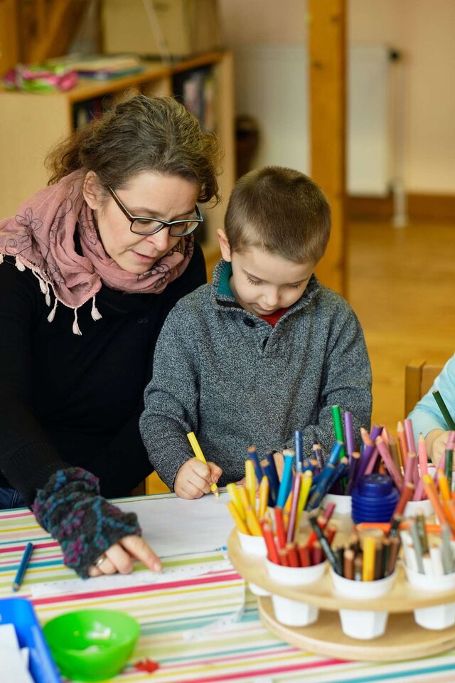 Eine Erzieherin sitzt mit einem Jungen an einem Tisch. Er malt, sie zeigt ihm etwas
