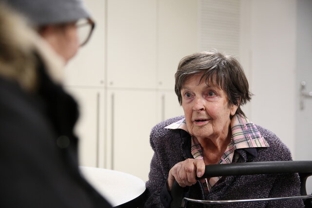 Annemarie Streit im Gespräch mit einer Besucherin des Mecki-Ladens. Man sieht die andere Person von hinten, Frau Streit schaut sie interessiert an.  