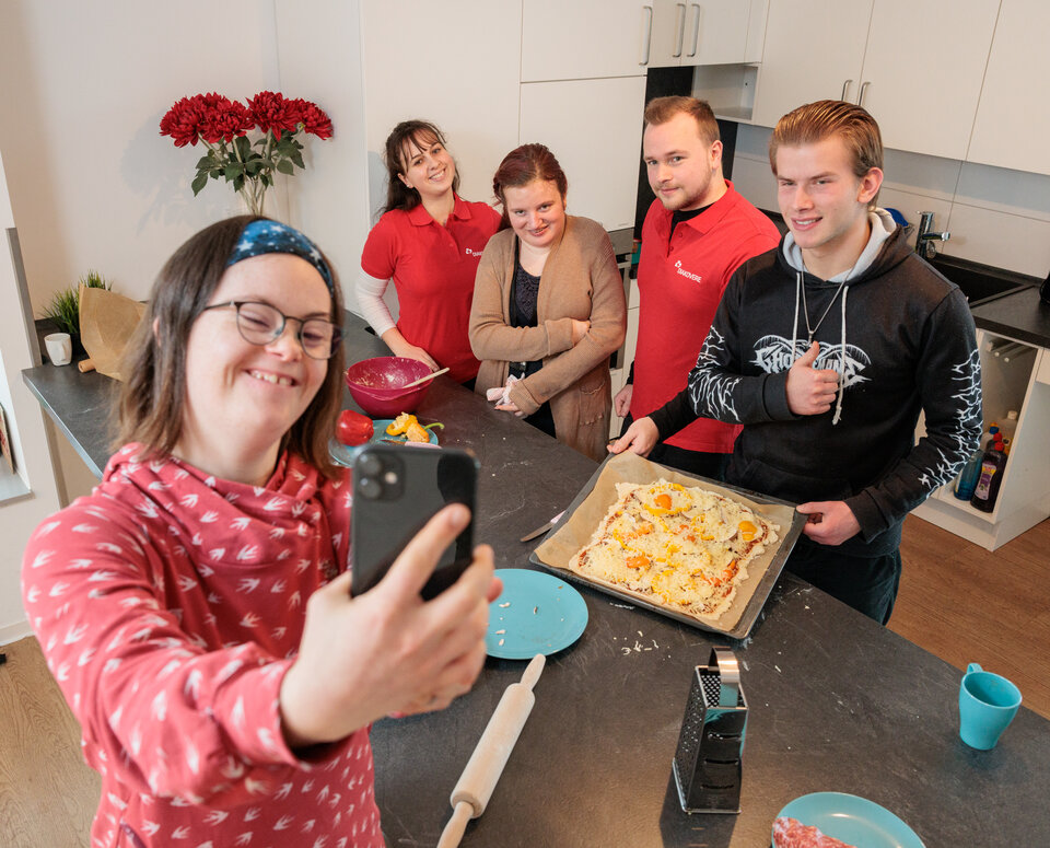 Fachverband Behindertenhilfe Eine junge Frau steht in der Küche ihrer Wohngruppe. Sie macht ein Selfie mit anderen jungen Menschen