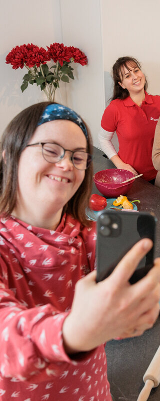 Fachverband Behindertenhilfe Eine junge Frau steht in der Küche ihrer Wohngruppe. Sie macht ein Selfie mit anderen jungen Menschen