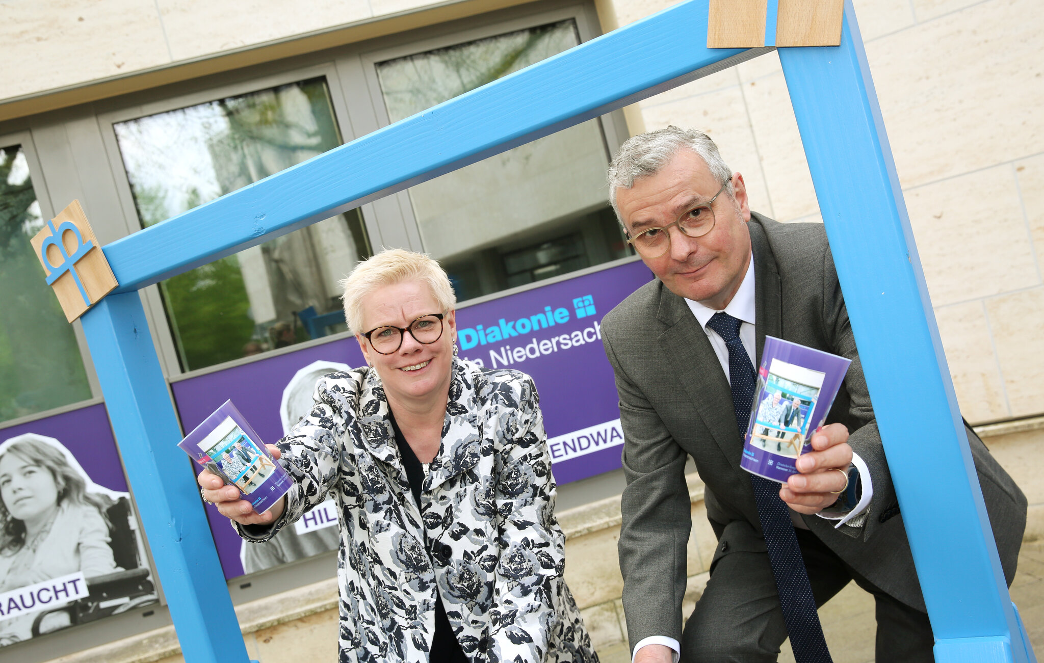 Zwei Personen (Marion Timm und Hans-Joachim Lenke) schauen durch einen mobilen Fotorahmen. Sie schauen fröhlich in die Kamera und halten Diakonie-Becher in der Hand.