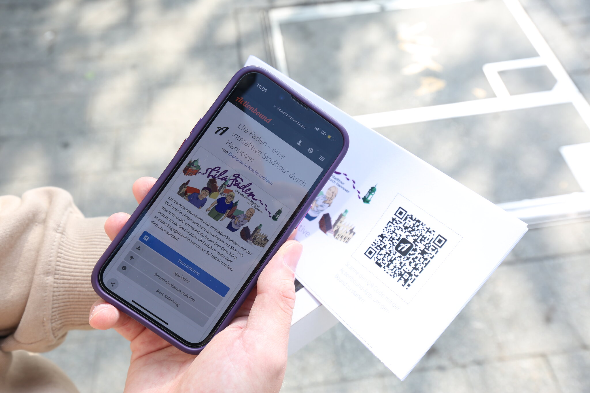 In der einen Hand hält eine Person ein Handy, in der anderen einen Flyer mit einem QR-Code. Sie scannt mit dem Handy den QR-Code. Von der Person sind nur die Hände zu sehen.