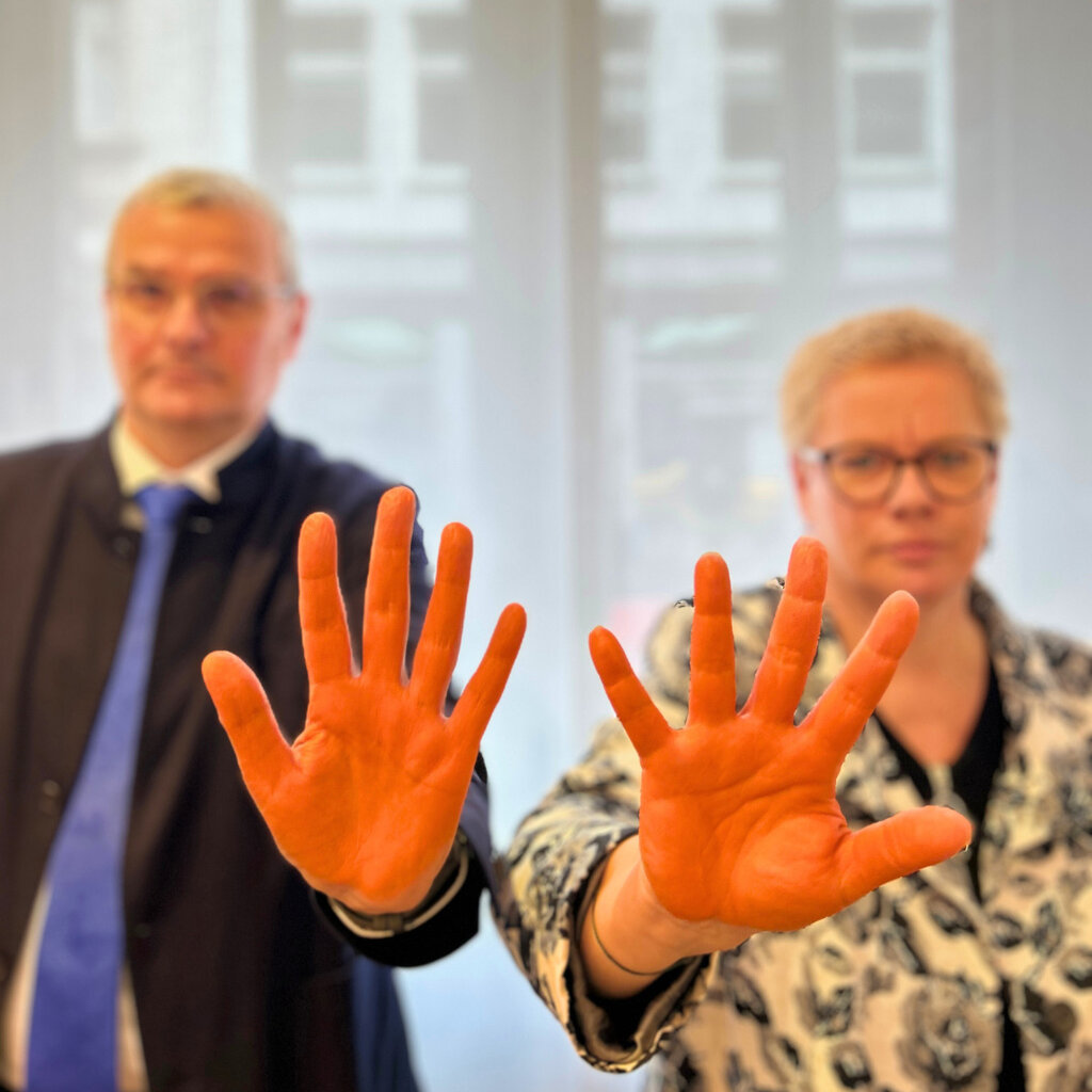 Die Vorstände Hans-Joachim Lenke und Marion Timm stehen nebeneinander, sie beide halten ihre orange-gefärbten Hände als "Stopzeichen" in die Kamera. Die Hände sind scharf, die Gesichter unscharf zu sehen