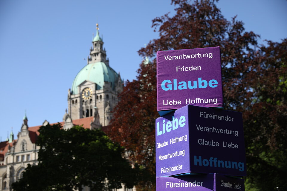 Diakonie-Würfel mit der Aufschrift Glaube, Liebe, Hoffnung stehen im Park. Dahinter ist das Hannoveraner Rathaus zu sehen
