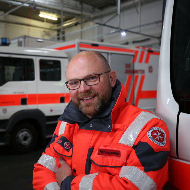 Ein Rettungssanitäter steht in seiner Uniform in der Halle mit Rettungswagen. Er schaut freundlich in die Kamera.