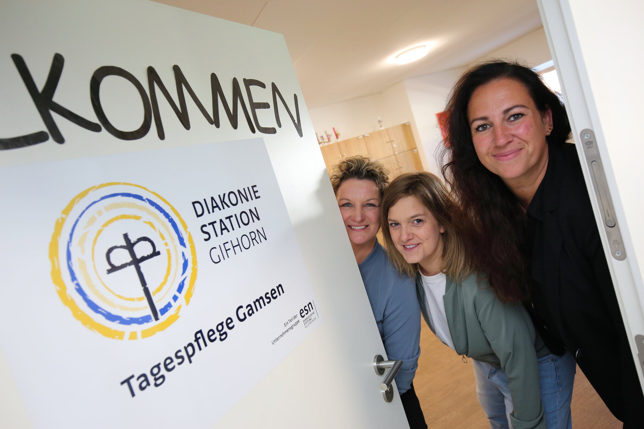 Birte Gessel, Bianca Pütsch und Janina Schoon stehen nebeneinander und schauen fröhlich in die Kamera. Neben ihnen steht ein Schild mit der Aufschrift "Willkommen"