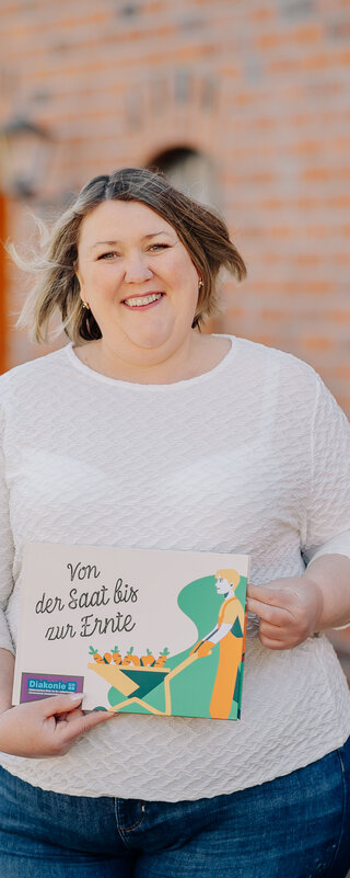 Eine Frau schaut fröhlich in die Kamera, in der Hand hält sie ein Bild mit der Aufschrift "Von der Saat bis zur Ernte"